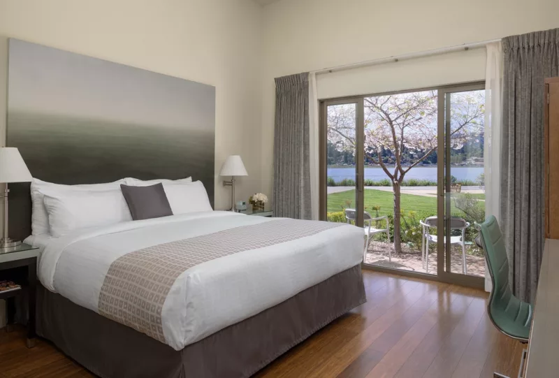 Hotel room with King bed, large art piece over bed, bedside tables and lamps. large sliding glass doors lead to patio with chairs and blooming cherry blossom tree