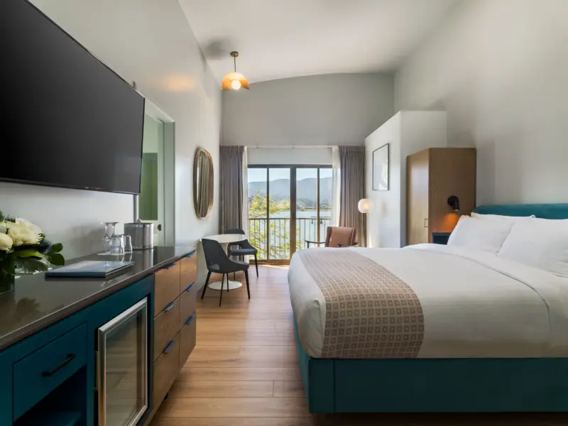 Hotel room with King bed, large art piece over bed, bedside tables and lamps. large sliding glass doors lead to patio with chairs and blooming cherry blossom tree
